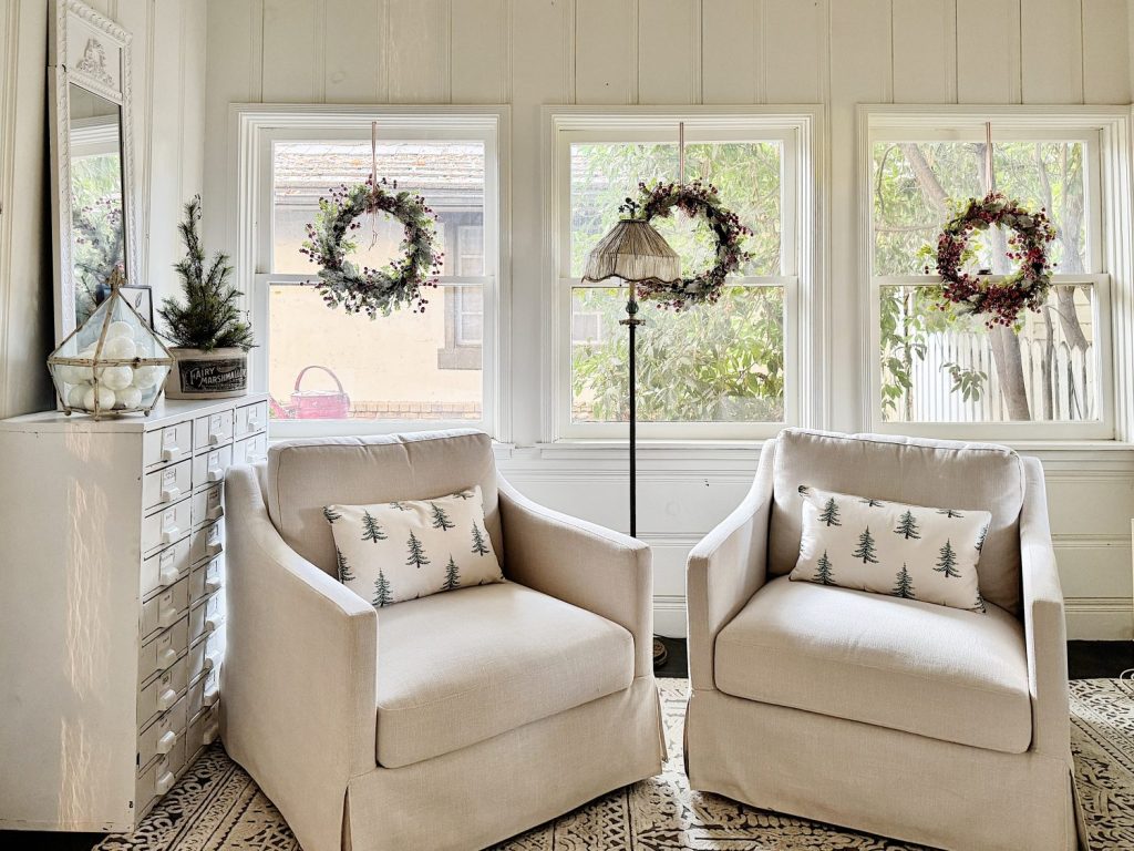 Two beige armchairs with tree-patterned pillows sit by windows decorated with hanging wreaths. A small tree and a glass lamp adorn a white dresser nearby.