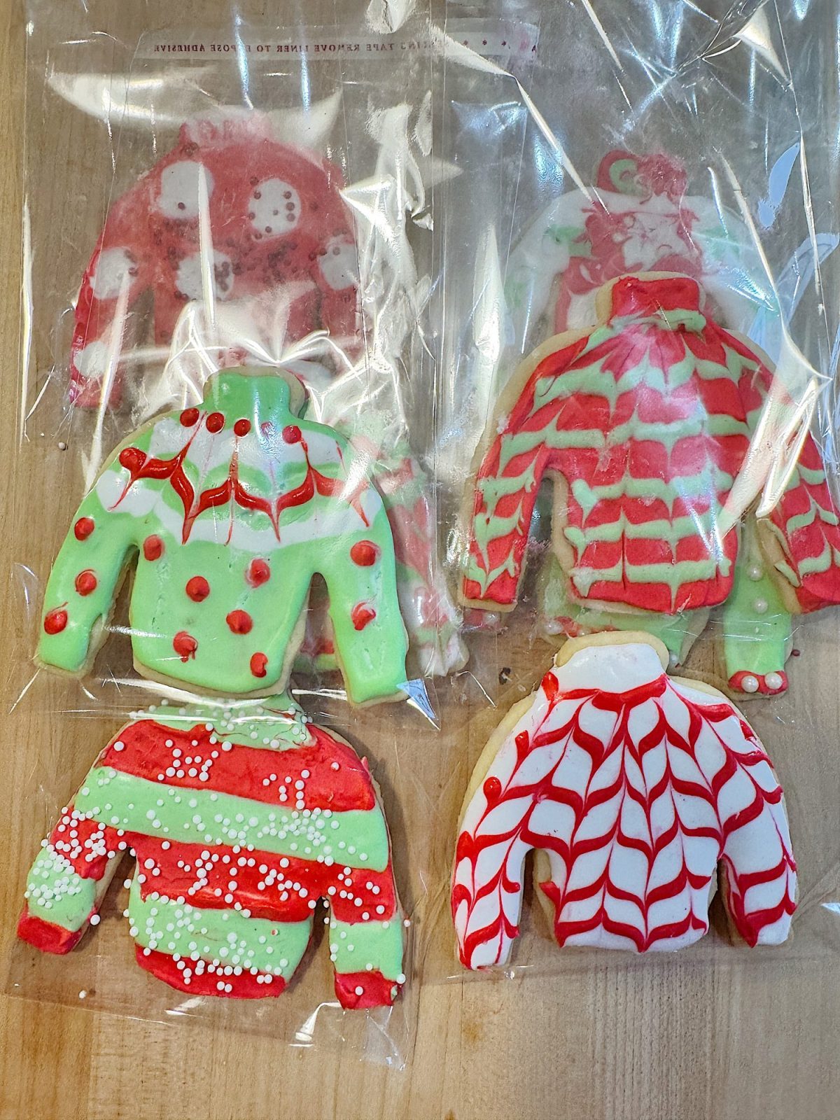 Four decorated sweater-shaped cookies with holiday designs in red, green, and white icing, packaged in clear plastic bags on a wooden surface.