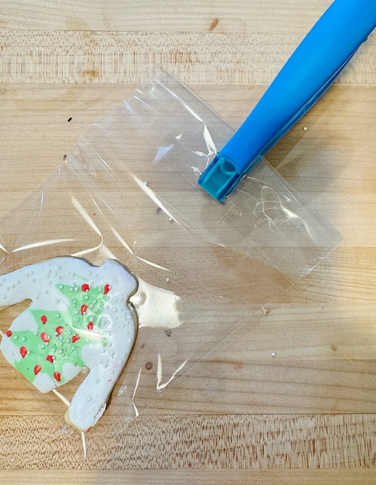 A frosted Christmas tree cookie is seen through a clear plastic bag on a wooden surface. A blue cookie scoop handle extends from the bag.