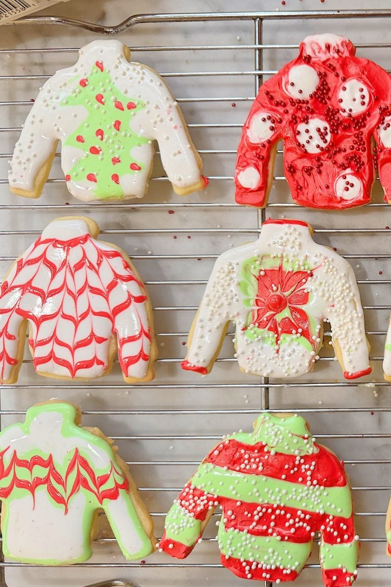 Six sweater-shaped cookies decorated with holiday-themed icing in various patterns, on a cooling rack.