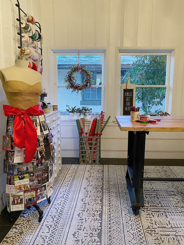 Craft room with a dress form adorned with a red ribbon, holiday cards, and a wreath hanging in the window. Gift wrap is stored in a corner.