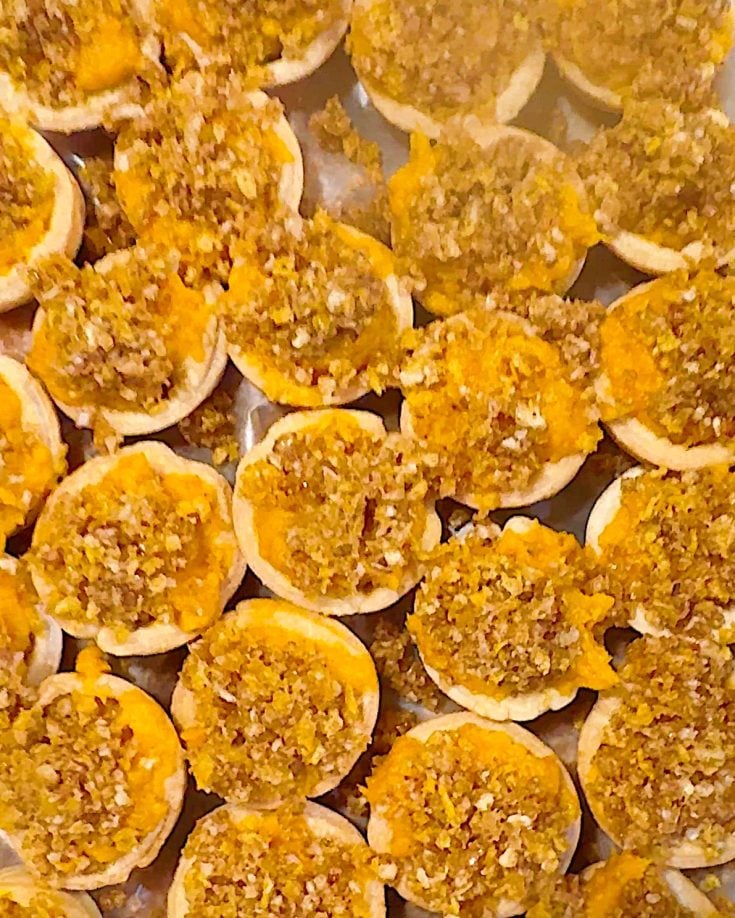 Close-up of multiple baked tarts, perfect for a Christmas party, filled with a crumbly, golden-brown topping and arranged in festive rows on a tray.