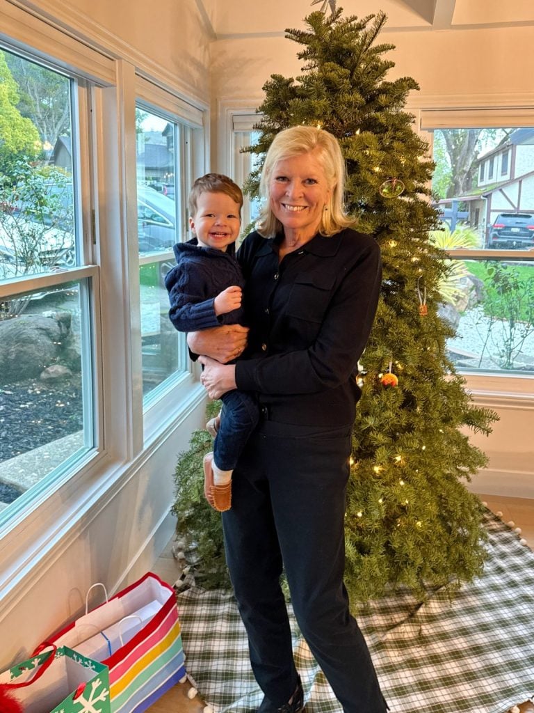 A woman holds a smiling child next to a decorated Christmas tree, with wrapped gifts on the floor nearby.