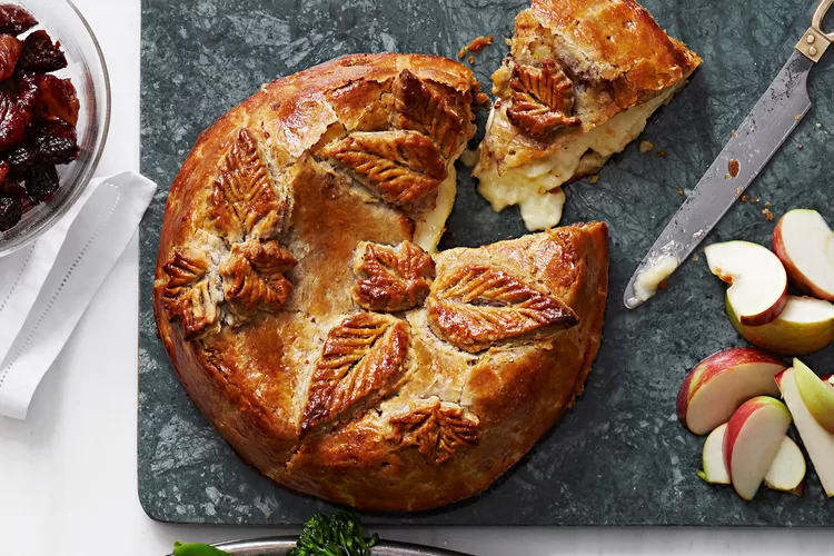 Baked Brie with decorative pastry leaves on a slate board, partially sliced, beside fresh apple wedges and a knife.