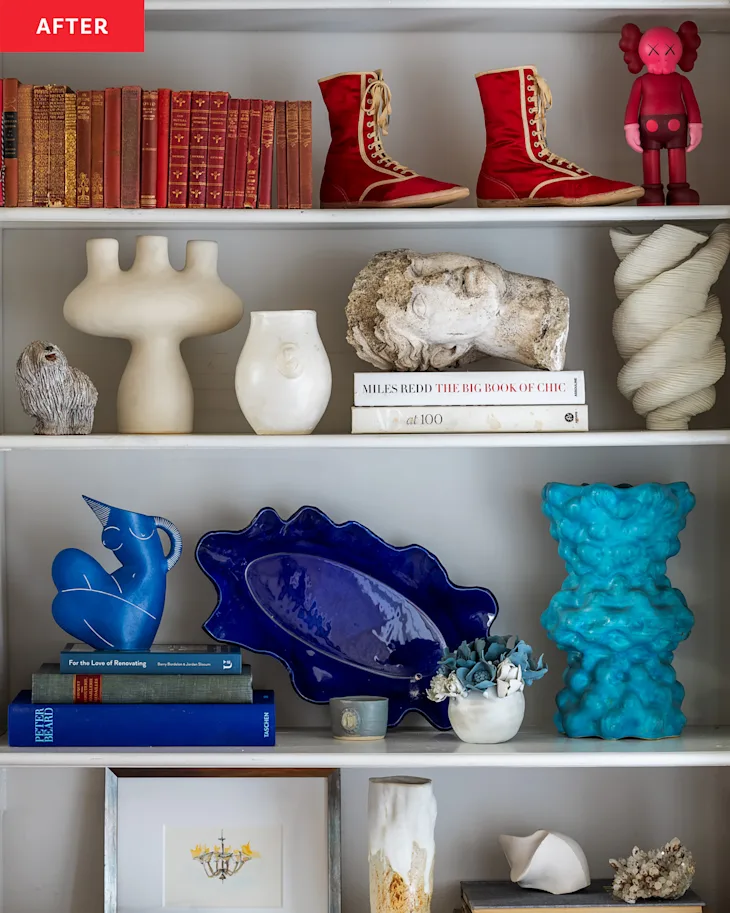 Bookshelf with decorative items: red shoes, books, abstract sculptures, a rock, vases, and a small figure. Blue and white pottery is on the lower shelf.