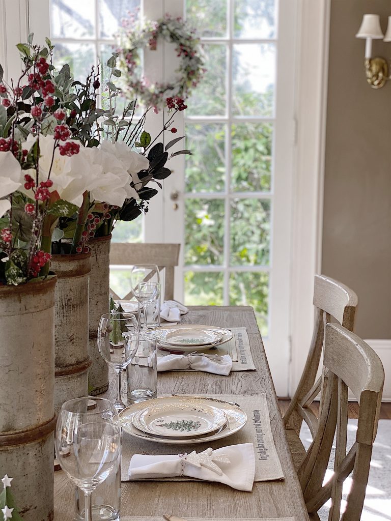 A dining table set for four with white plates, wine glasses, and napkins. Large floral arrangements in metal vases decorate the table. A wreath hangs on the glass door in the background.