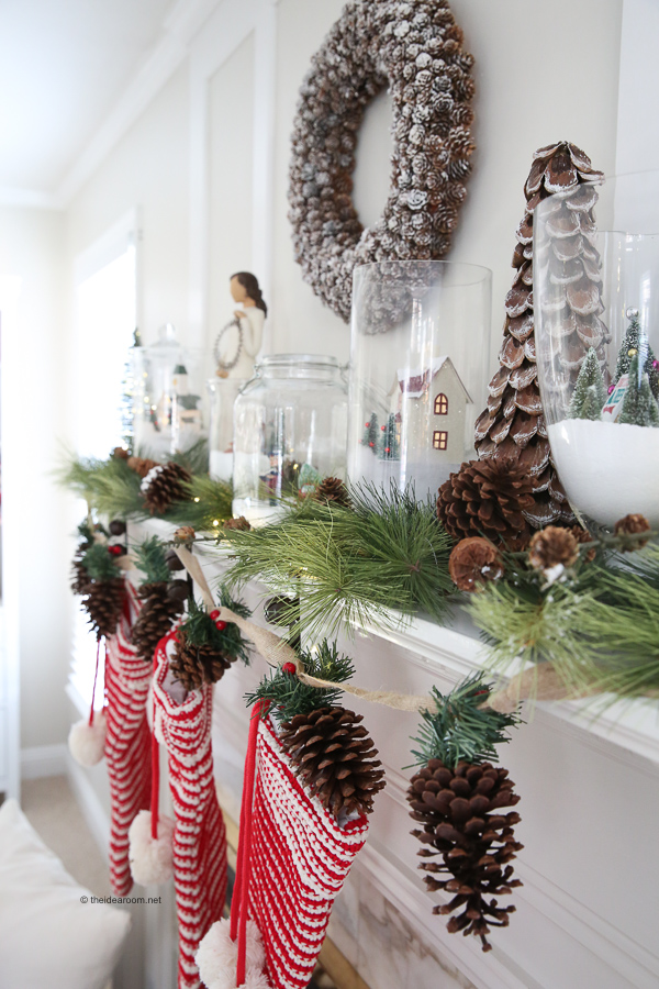 Christmas-themed mantel with stockings, pinecones, a wreath, and greenery. Decorative jars with snow and a small house sit on top, creating a festive winter scene.