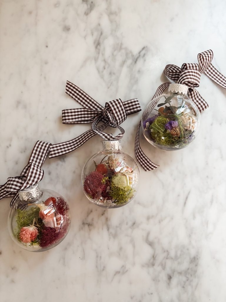 Three clear glass ornaments filled with colorful dried flowers and tied with checkered ribbons on a marble surface.