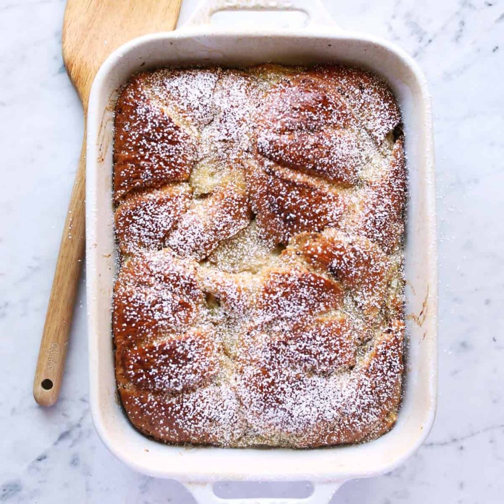 A baked dish of bread pudding topped with powdered sugar in a rectangular white baking dish. A wooden spatula rests beside it on a marble surface.