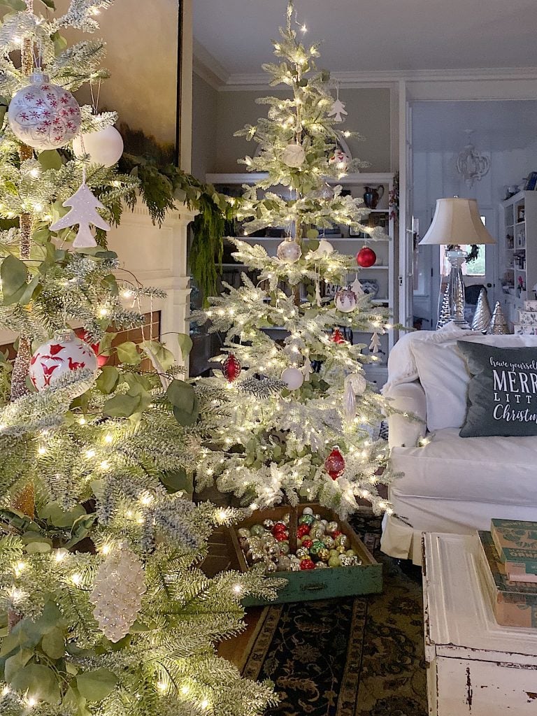 A living room with lit Christmas trees decorated with ornaments. A white sofa with a festive pillow is on the right, and a tray of additional ornaments is on the floor.