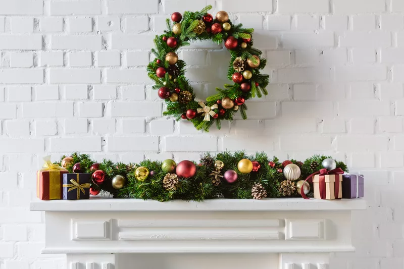 A Christmas wreath hangs on a white brick wall above a mantel decorated with garland, ornaments, pine cones, and gift boxes.