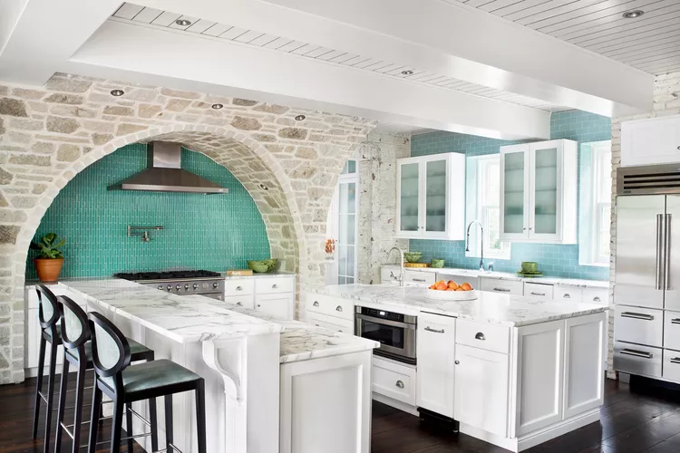 Modern kitchen with a curved stone arch, turquoise tile backsplash, white cabinetry, marble countertops, bar stools, and a stainless steel range hood and refrigerator.