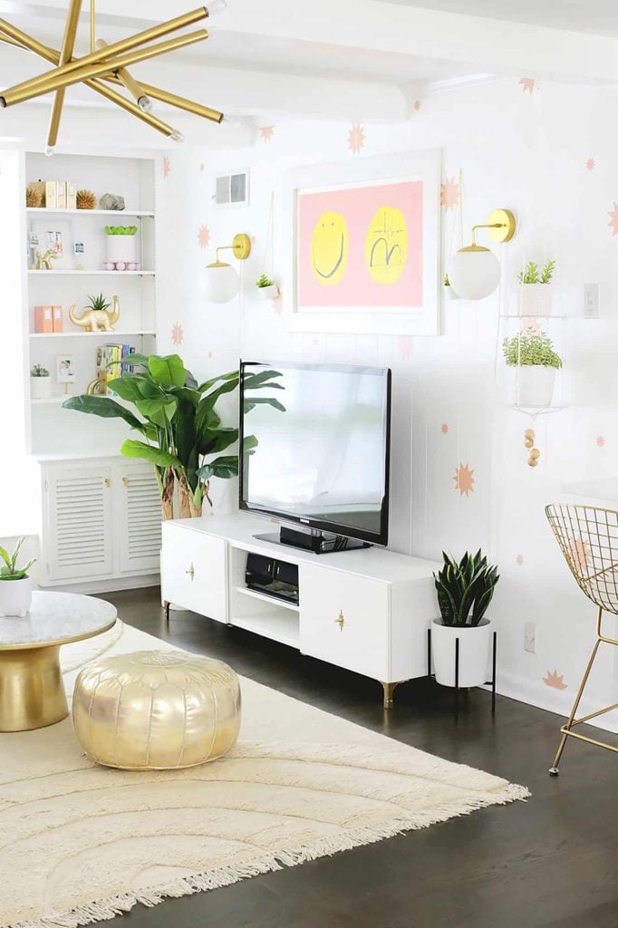 A modern living room with a white TV stand, wall-mounted TV, plants, and decorative wall art. A gold pouf and a geometric chandelier complement the minimalist design.