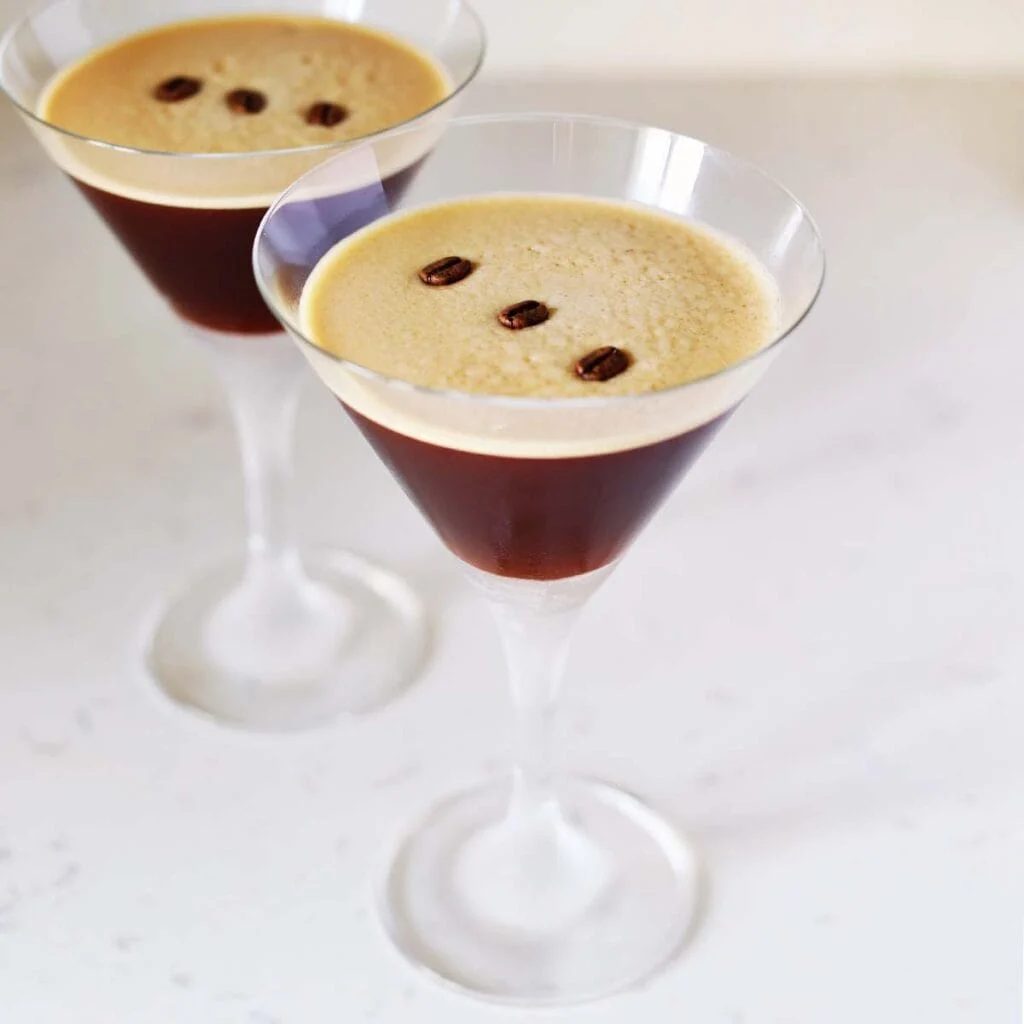 Two espresso martinis in cocktail glasses, each topped with three coffee beans, on a white surface.