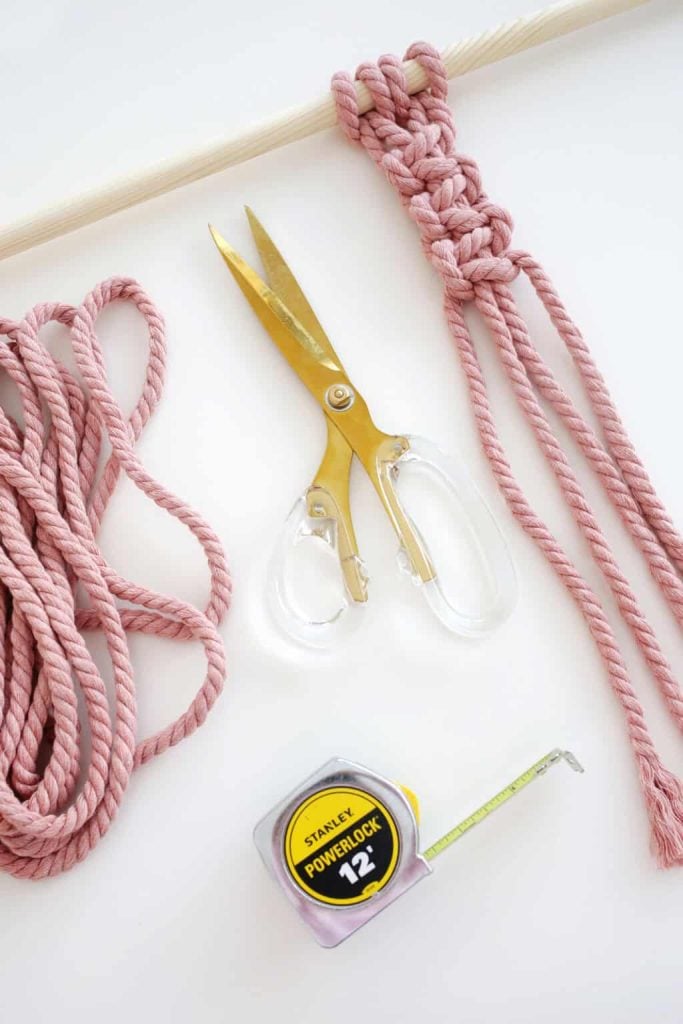 Pink macramé in progress with a wooden dowel, gold-handled scissors, pink rope, and a 12-foot tape measure on a white surface.