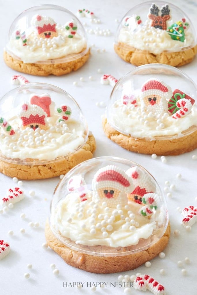 Cookies topped with festive decorations under clear domes, surrounded by white sprinkles on a white surface.