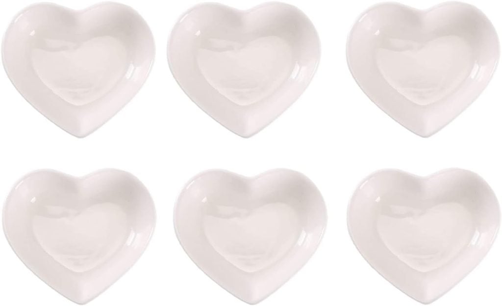 Six white, heart-shaped ceramic plates arranged in two rows of three on a plain background, perfect for creating a charming Valentine's Day charcuterie board.