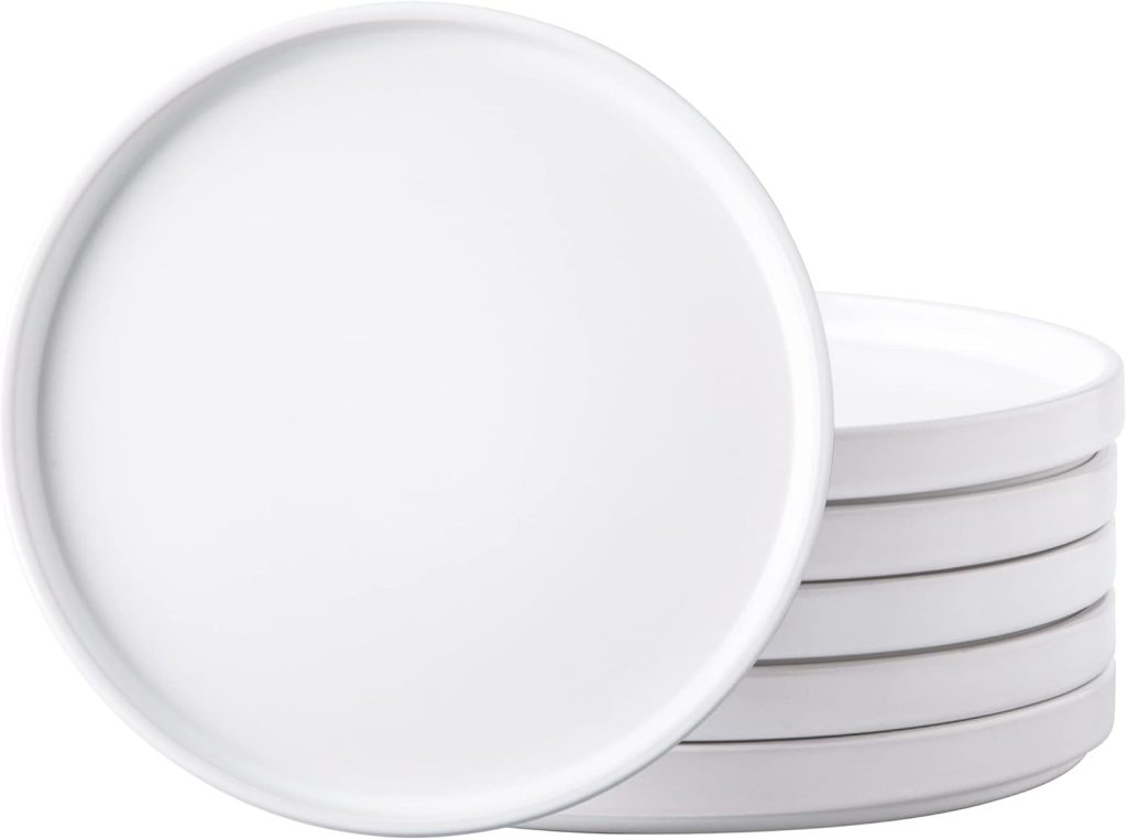 A stack of six plain white round ceramic plates, classic kitchen items, with one plate leaning against the stack.