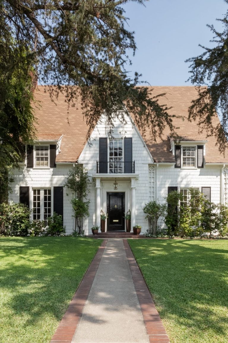 A white two-story house with a brown roof, black shutters, and a central door. It has a manicured lawn and a red brick pathway leading to the entrance. Trees frame the scene.