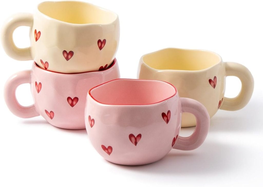 Four ceramic mugs, two pink and two cream, each decorated with small red hearts, are stacked on a white background&mdash;perfect for serving drinks alongside your Valentine's Day Charcuterie Board.