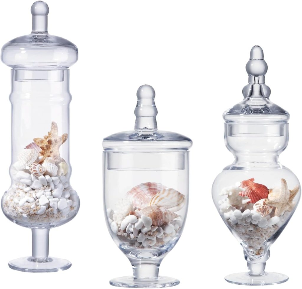 Three clear glass jars with lids, each containing a mix of seashells and small white stones&mdash;arranged side by side against a white background&mdash;evoke the charm of displaying Valentine&rsquo;s Day sugar cookies in decorative containers.