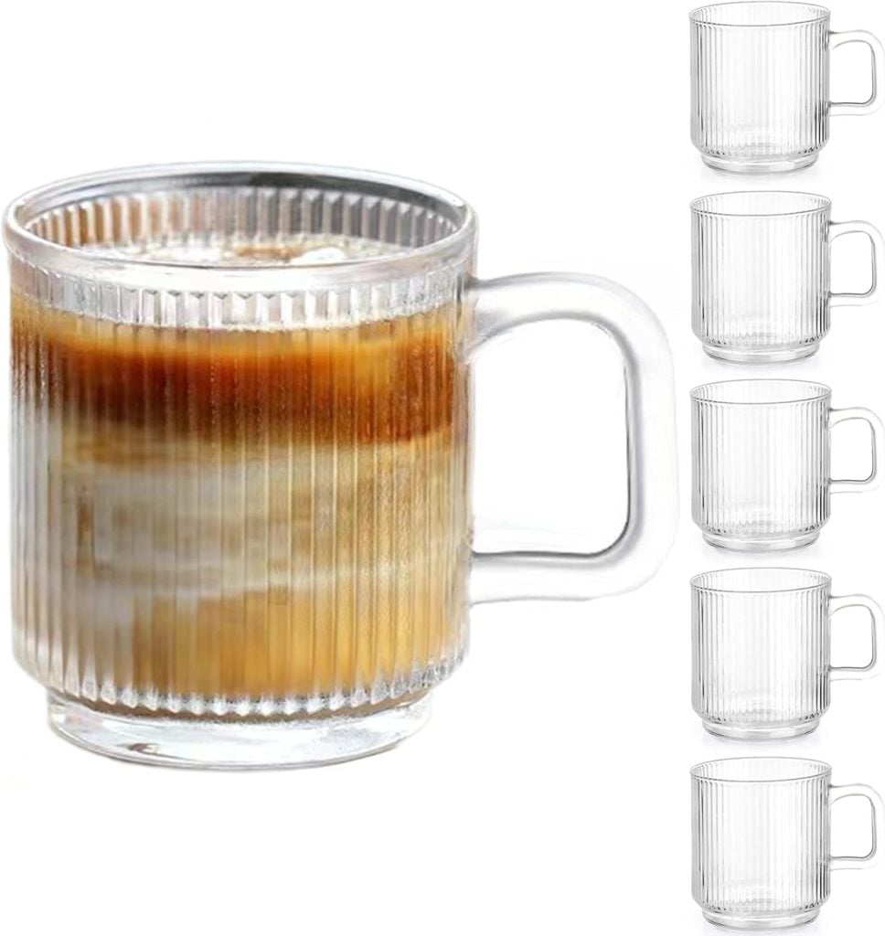 A transparent glass mug filled with layered coffee and milk is displayed on open kitchen shelving next to an evenly spaced stack of four identical empty mugs.