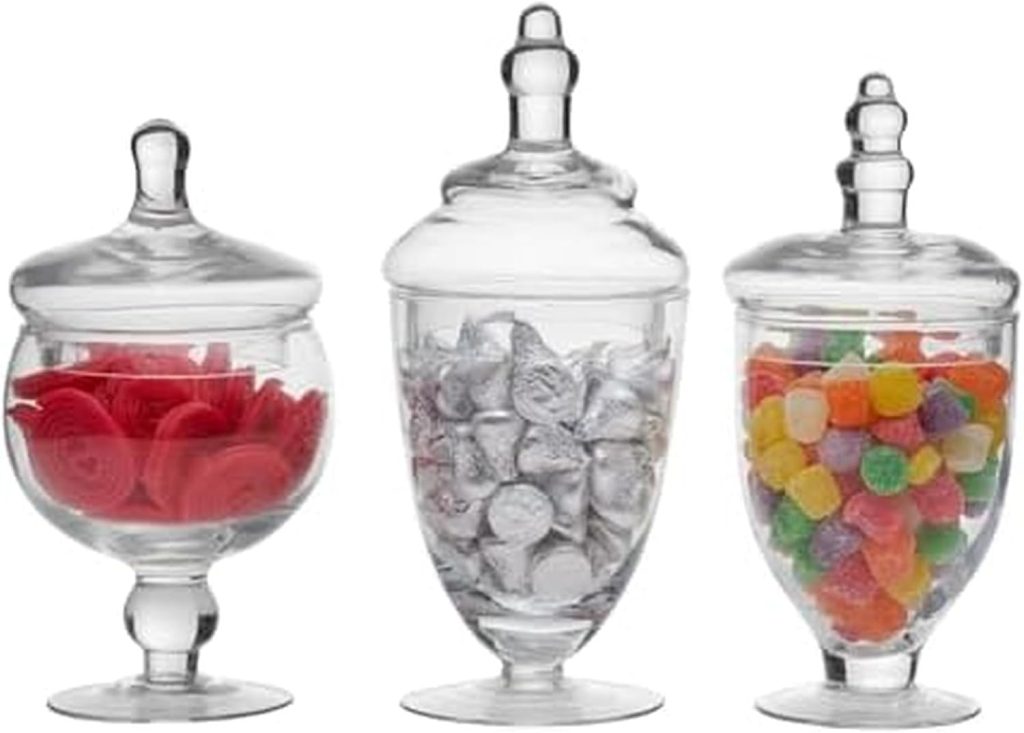 Three glass apothecary jars with lids, each filled with different candies: red candies on the left, silver-wrapped chocolates in the middle, and assorted gumdrops on the right.