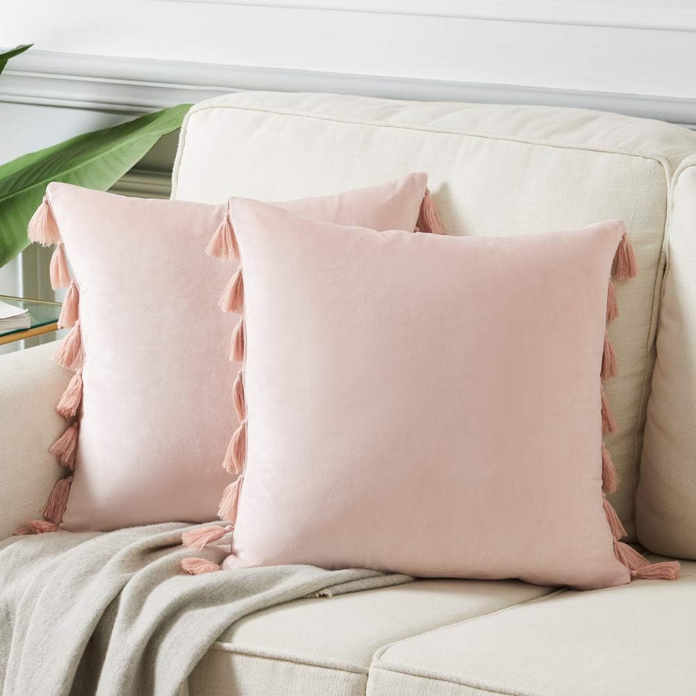 Two light pink square pillows with tassels on the edges are placed on a light beige sofa next to a heart pillow and a light gray blanket.