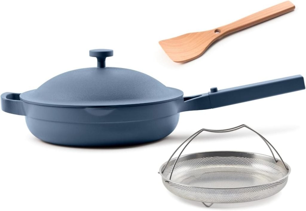 A blue nonstick pan with a lid and handle, accompanied by essential kitchen items like a wooden spatula and a metal steamer basket, is artfully arranged against a white background.