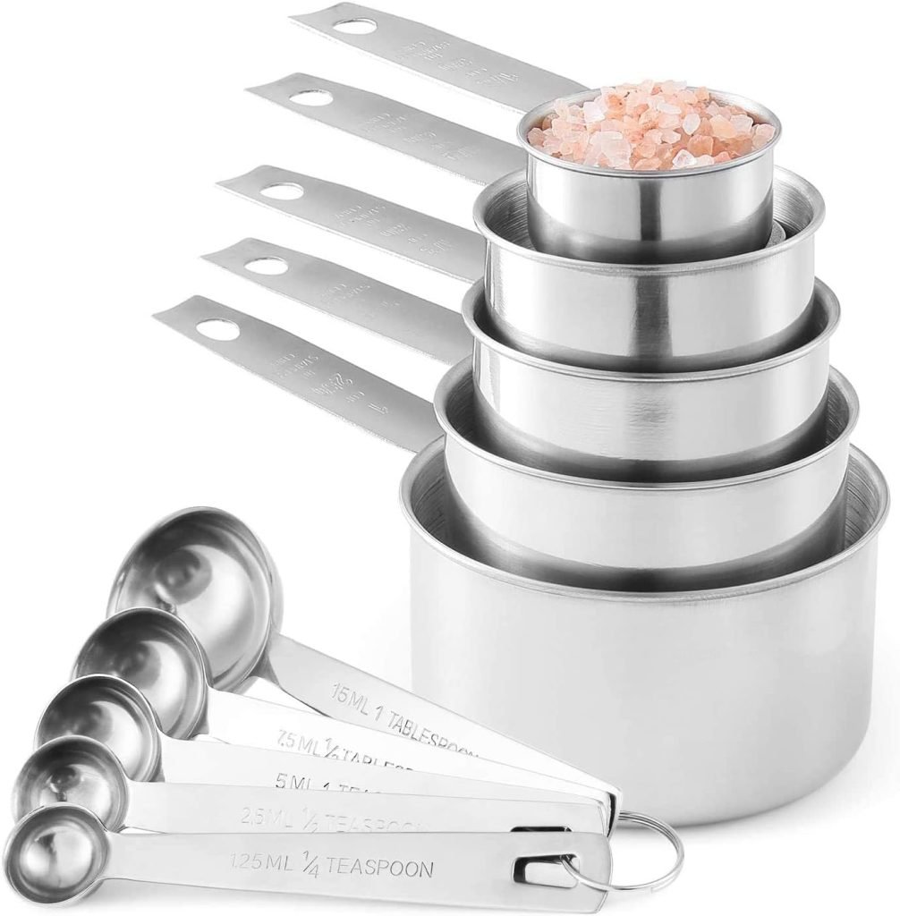 A set of stainless steel measuring cups and spoons, with one cup holding pink salt, while a sprinkle of brown sugar rests nearby.