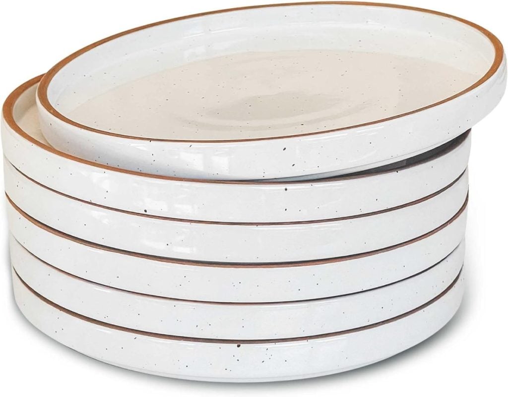 A stack of six white ceramic plates with brown edges and speckled patterns sits elegantly on the open kitchen shelving, adding a touch of rustic charm to the display.