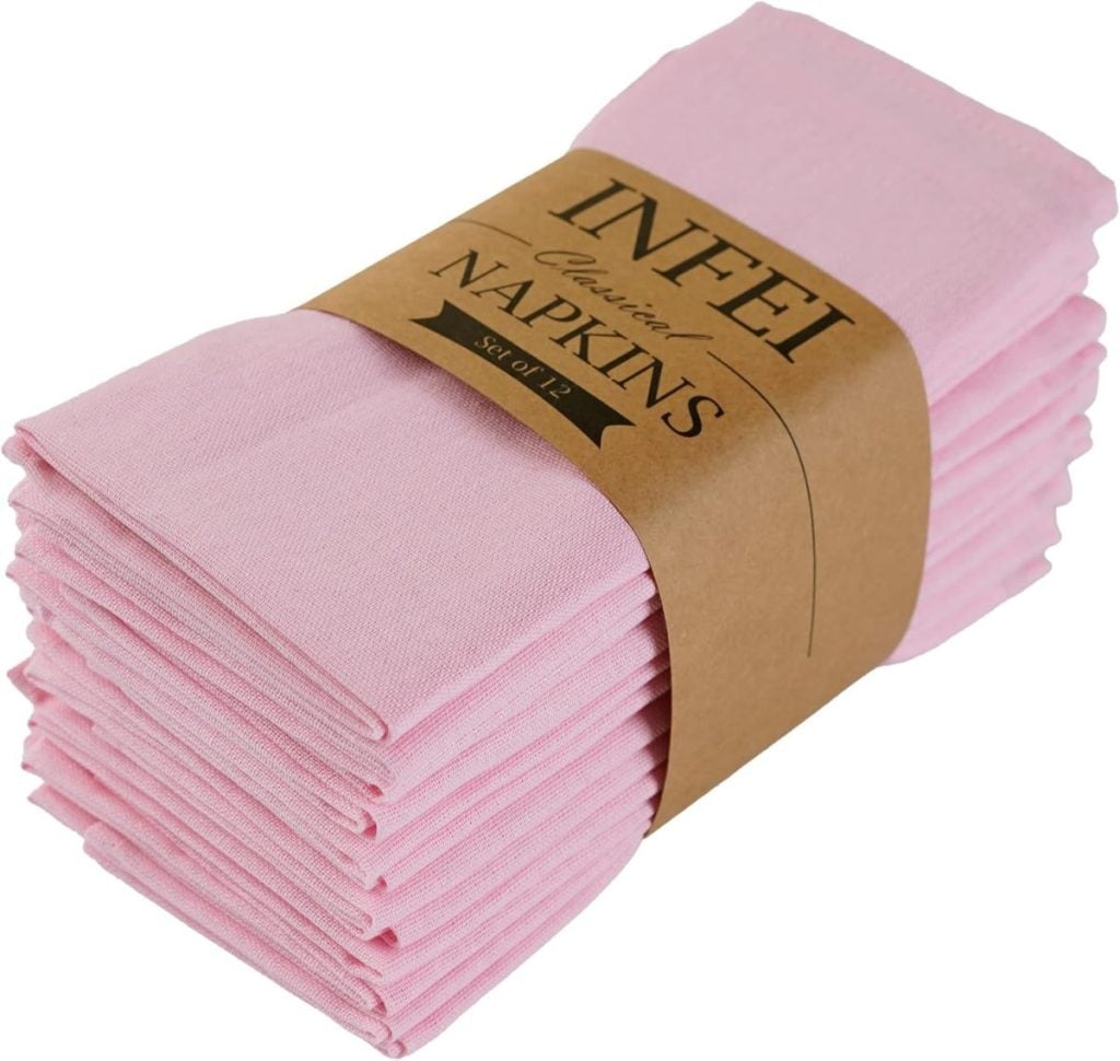A stack of folded pink cloth napkins wrapped with a brown paper band labeled "INFEI Classic Napkins Set of 12," perfect for Valentine table decorations.