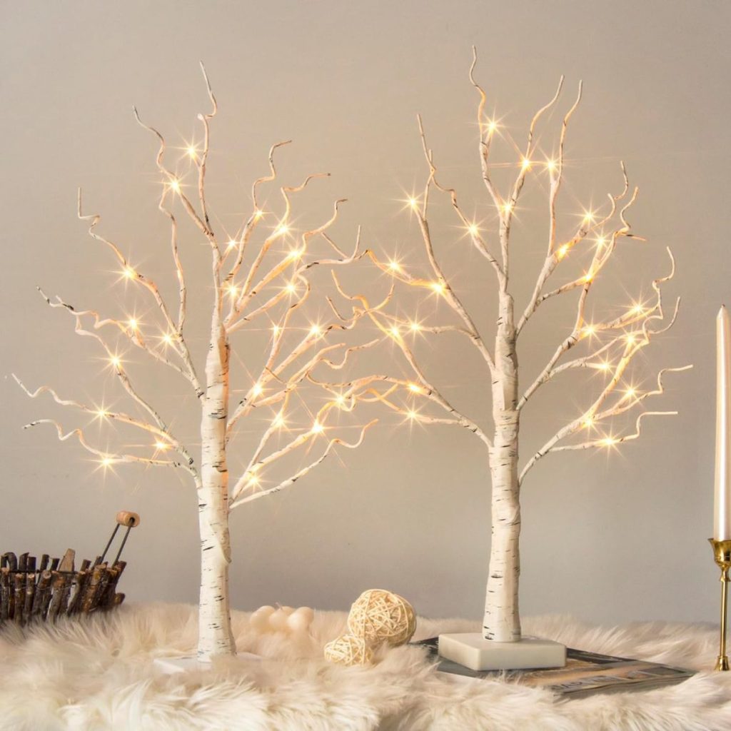 Two decorative LED birch trees with lights on a soft surface, accompanied by a candle and woven balls, creating a warm, cozy atmosphere.