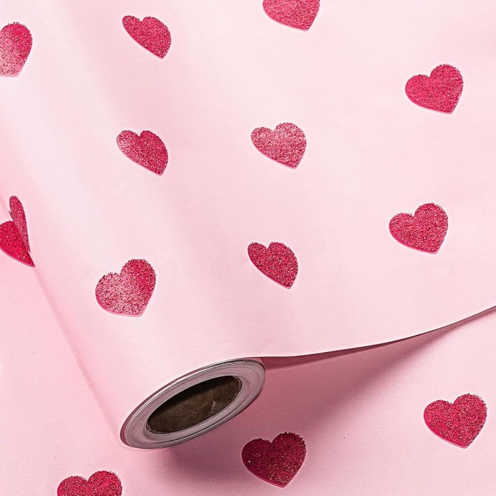 A roll of pink wrapping paper with a repeating pattern of red hearts, perfect for gifts or lining glass apothecary jars to add a sweet, romantic touch.
