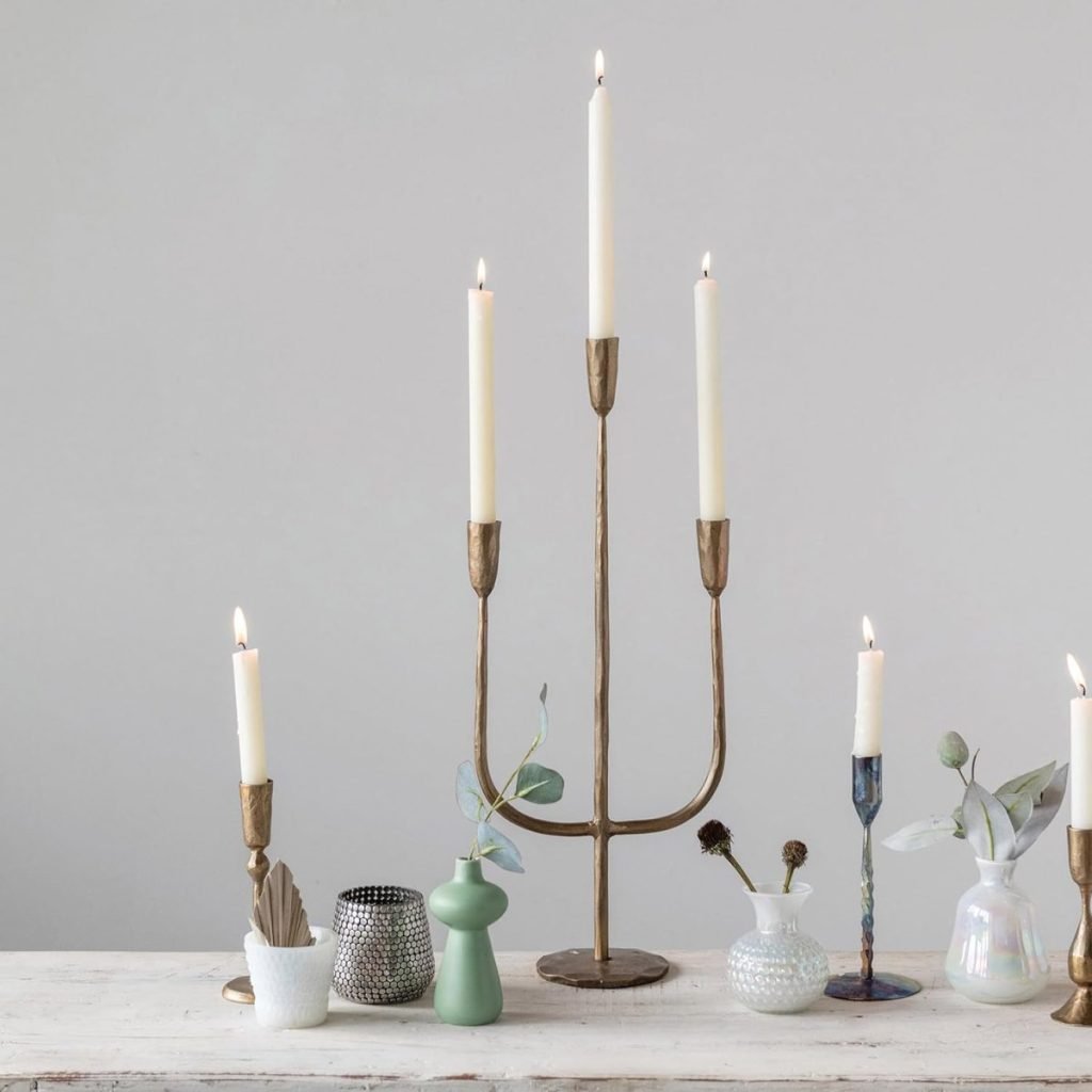 A brass candelabra with lit candles is surrounded by small vases and candle holders on a white surface against a neutral wall.