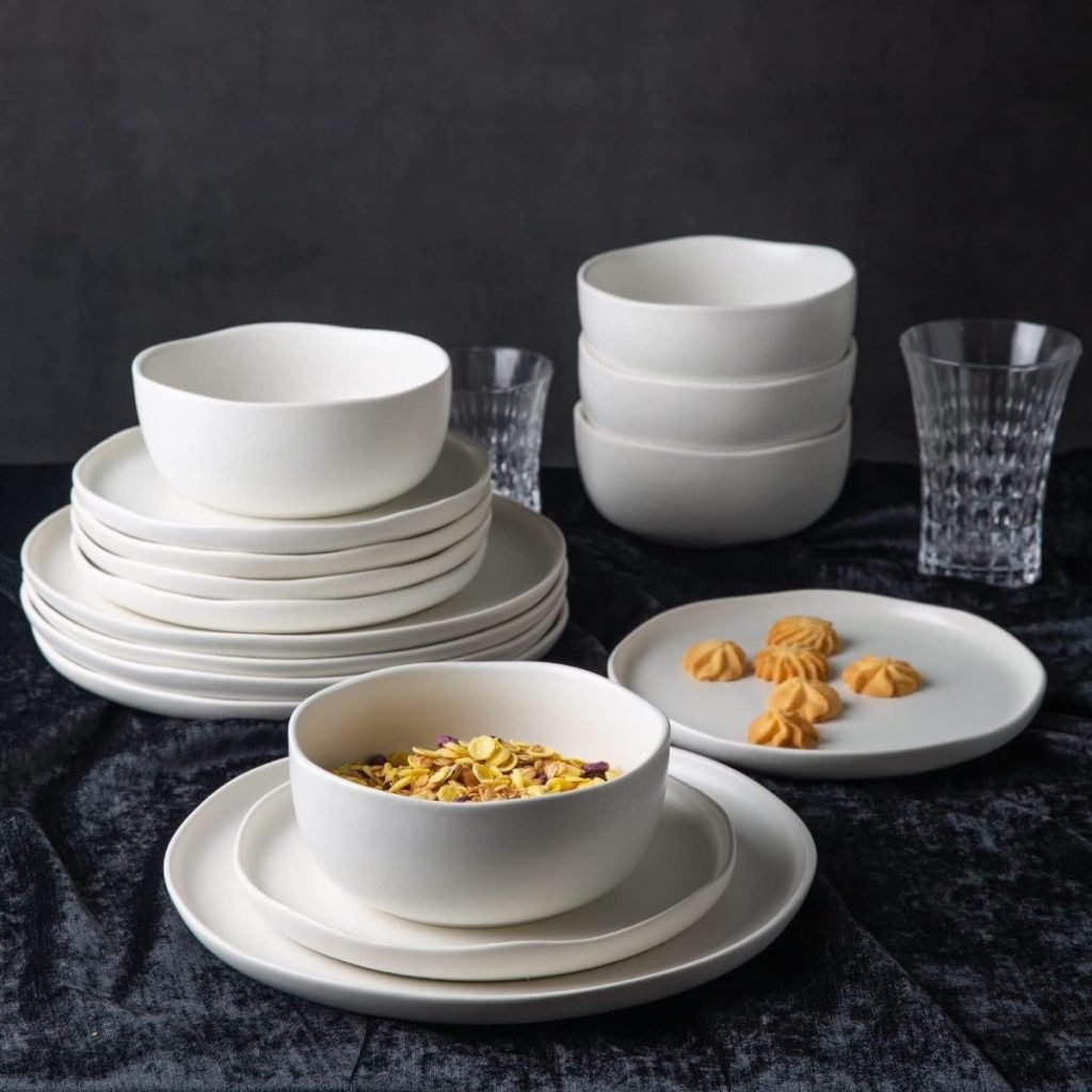 On dark cloth, a stack of white ceramic plates and bowls sits elegantly beneath open kitchen shelving. A bowl cradles cereal, while a plate presents five small cookies beside two clear glasses.