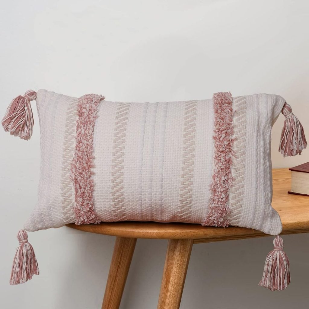 A rectangular, textured white cushion with pink tassels on each corner and two pink fringed stripes, reminiscent of a Heart Pillow, placed on a wooden chair next to a closed book.