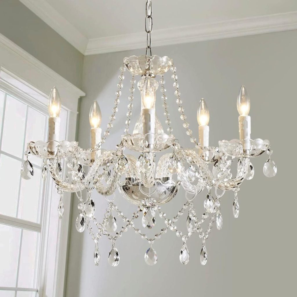 Crystal chandelier with six lit candle-style bulbs, glass beads, and hanging crystal pendants, suspended above Valentine table decorations in a room with light gray walls and a large window.