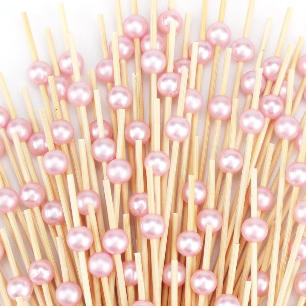 A cluster of wooden sticks, each topped with a shiny, pink pearl-like bead resembling chocolate marshmallow treats, arranged closely together on a white background.