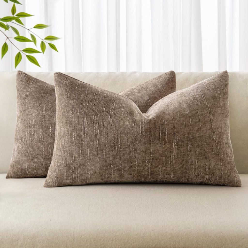 Two beige textured throw pillows on a light-colored couch with a leafy branch and sheer curtains in the background.