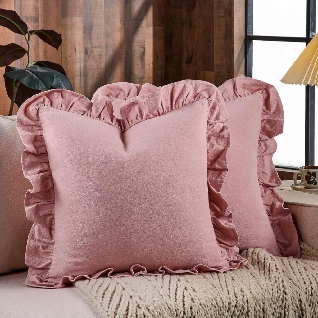 Two pink square pillows with ruffled edges sit side by side on a beige sofa, accompanied by a charming heart pillow, in a room with wooden walls and large windows.