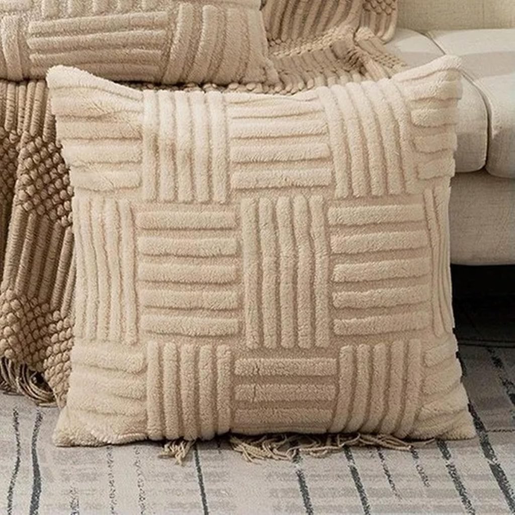 A beige textured throw pillow with a geometric pattern sits on a striped rug next to a matching woven blanket and a light-colored sofa.