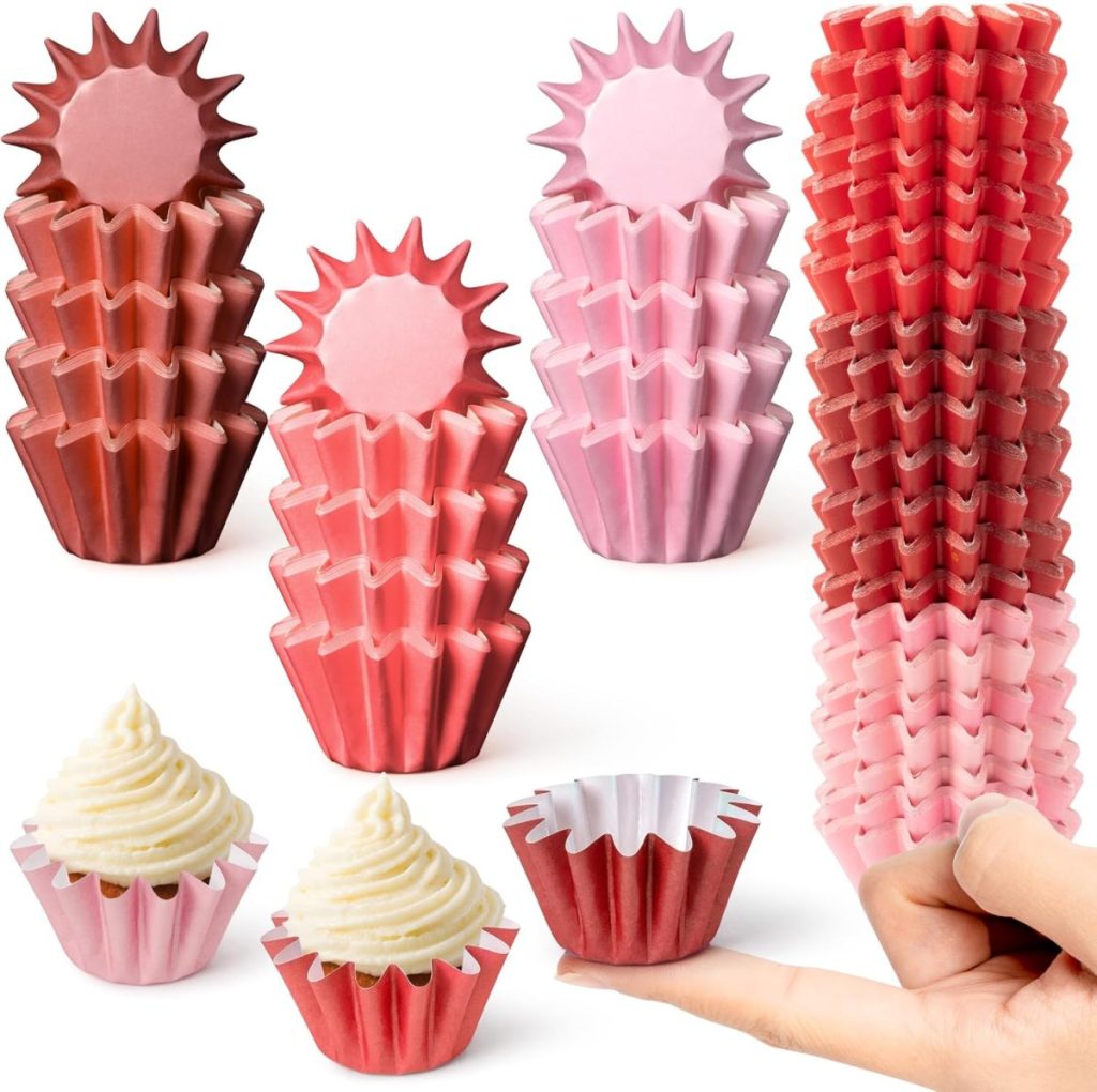 Stacks of pink and red cupcake liners in various shades, with two Valentine's cupcakes topped with frosting and a hand holding one liner in the foreground.
