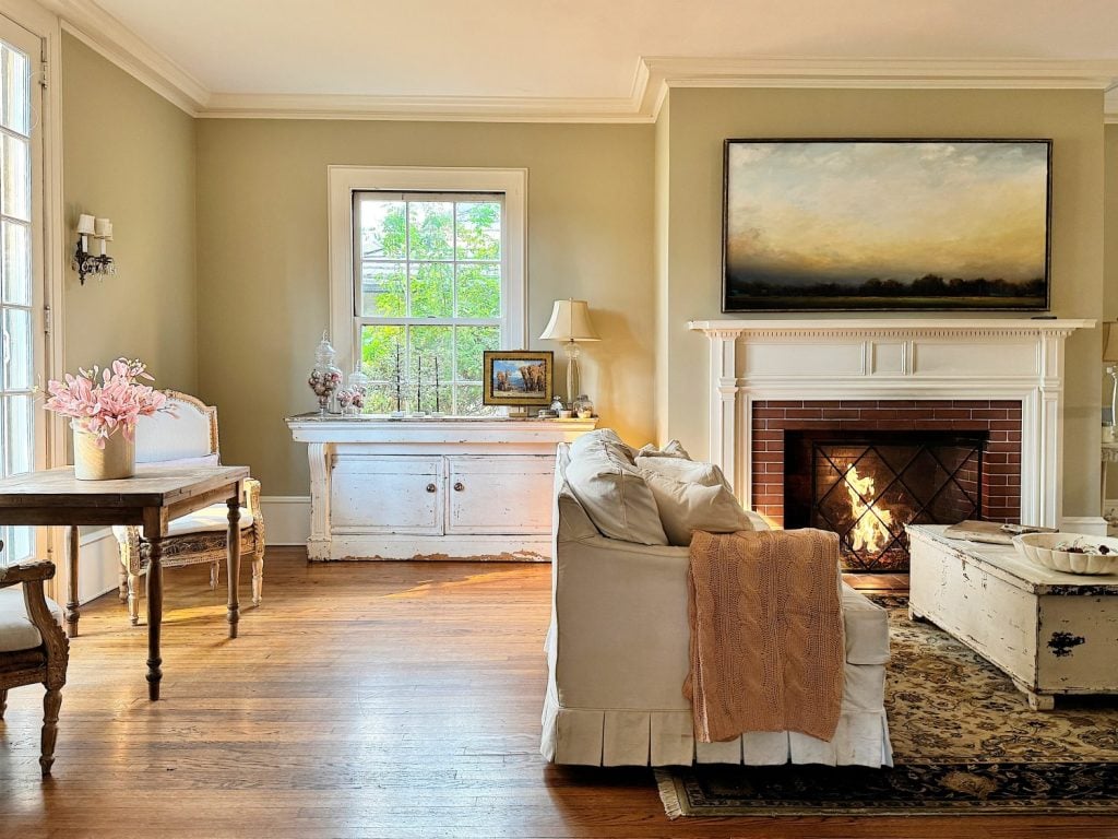 Cozy living room with a lit fireplace, white sofa, and light wood floor. A large window illuminates the room. A table with pink flowers is positioned near the window.