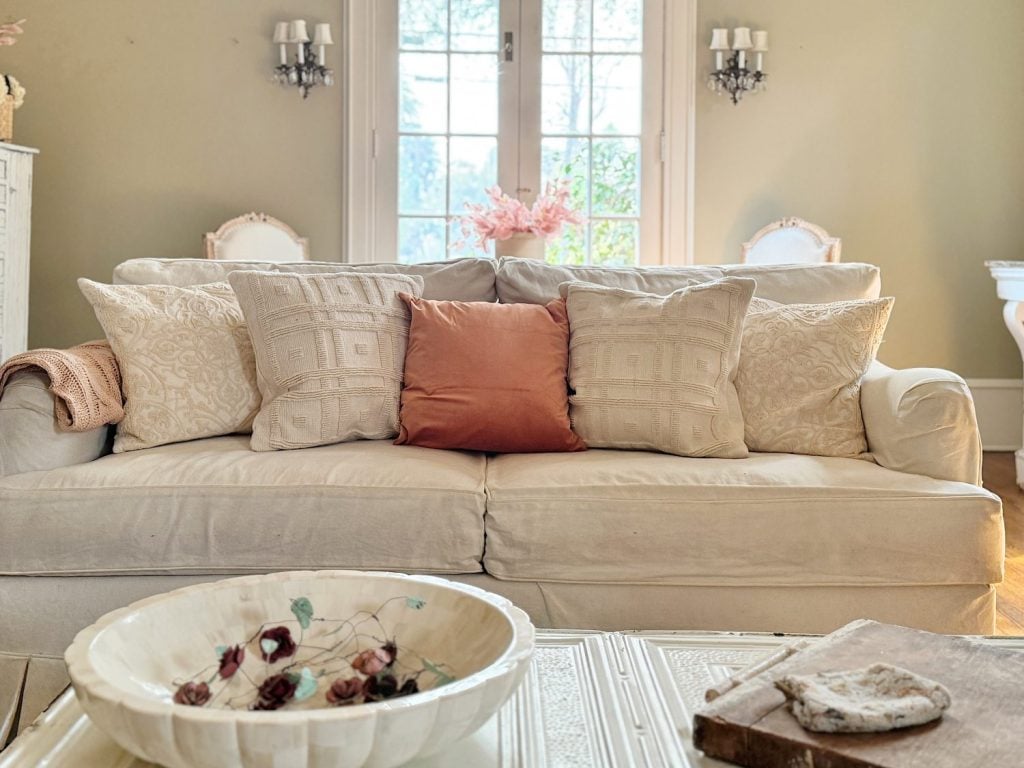 A cozy sofa with five decorative pillows, including a standout rust-colored one, sits in a well-lit living room with a large window and elegant decor. A floral bowl is on the coffee table.