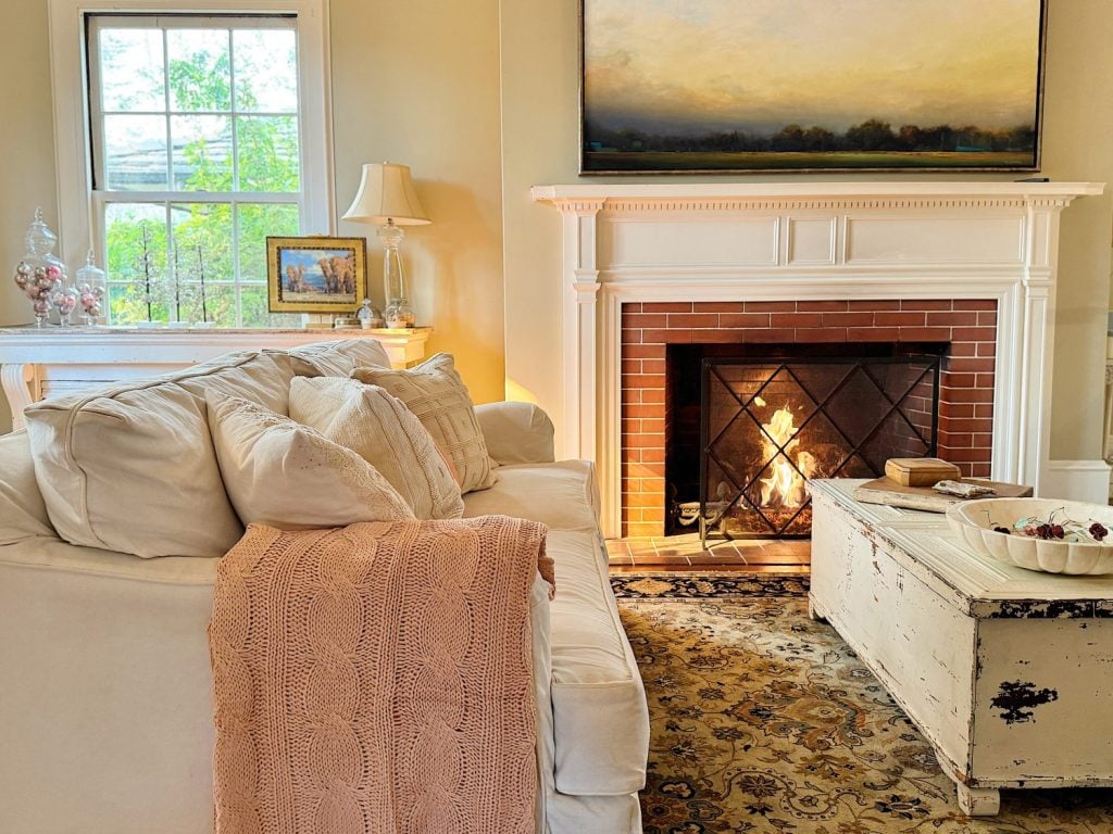 Cozy living room with a white sofa, pink blanket, lit fireplace, and a rustic coffee table. Large landscape painting above the mantel and a window letting in natural light.