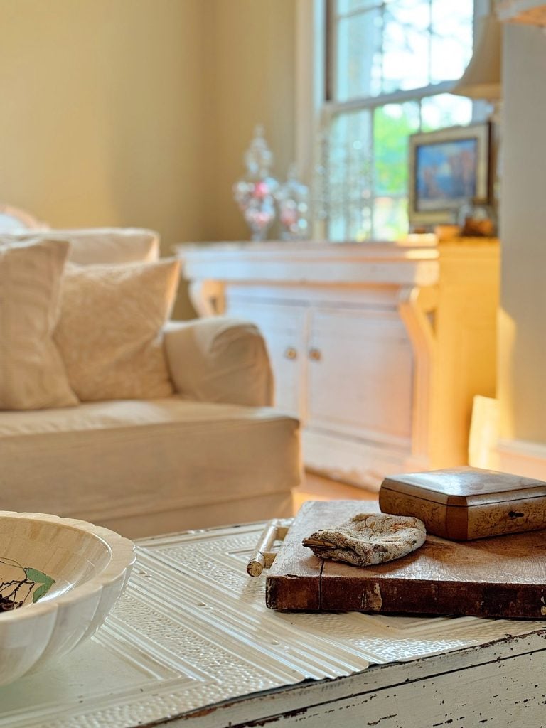 Cozy living room with a beige sofa, white sideboard, and a coffee table. The table has a bowl, wooden box, and a piece of driftwood. Sunlight filters through a window in the background.