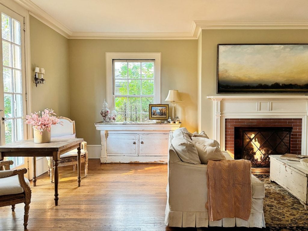 A cozy living room with a lit fireplace, a beige sofa, a wooden table with chairs, and a window overlooking greenery. A decorative painting hangs above the mantel.