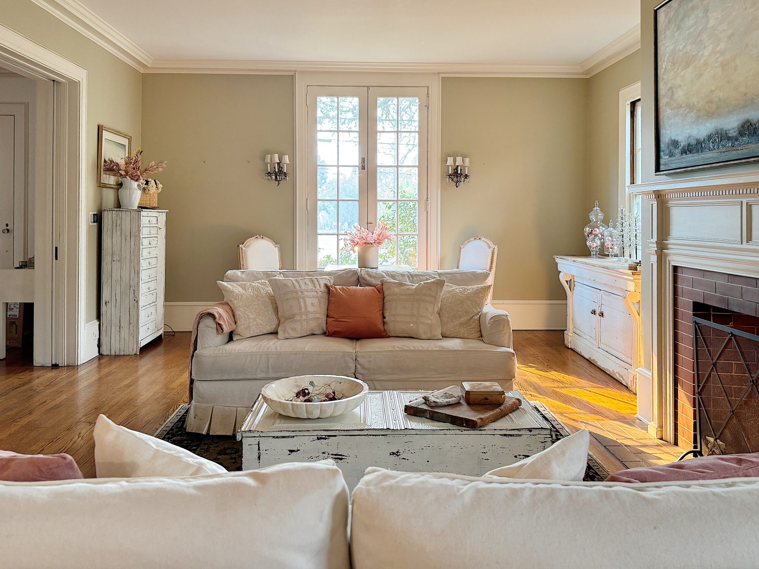 A cozy living room with a cream sofa, orange cushions, and light wooden furniture. Sunlight filters through tall windows, highlighting decorative vases and a painting above the fireplace.