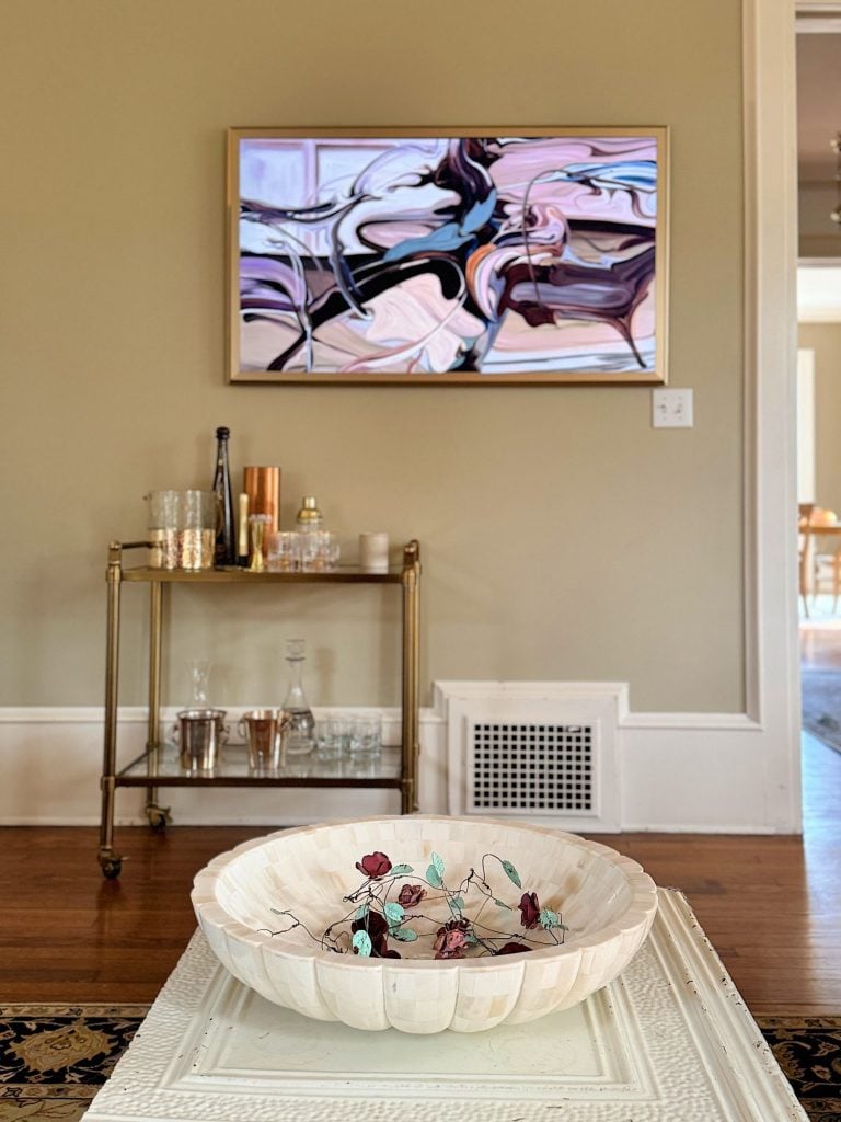 A living room features a wall-mounted abstract painting and a bar cart with glassware. A decorative bowl with colored leaves is on the table in the foreground.